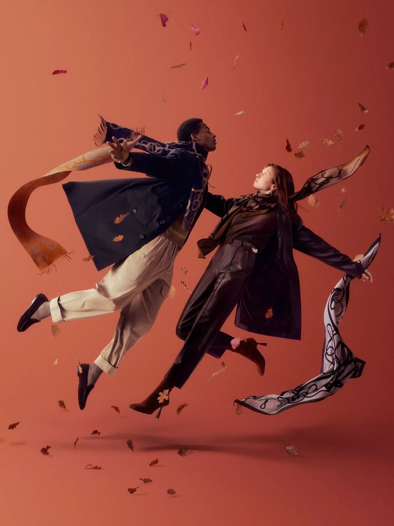 Two fashion models in mid-air wearing coats, trousers, and scarves, surrounded by falling leaves against a red-orange studio backdrop.