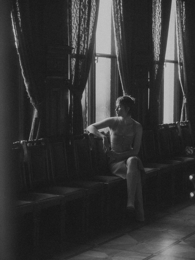 Monochrome portrait of a woman sitting on a cushioned bench in front of a window with patterned curtains in an indoor setting.