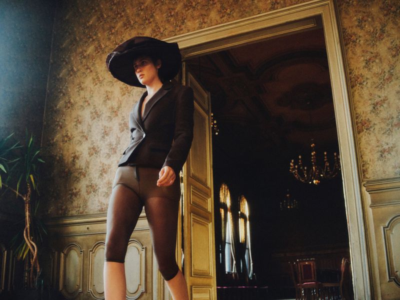 A woman in a dark suit and large hat walks through a doorway into a dimly lit room with ornate wallpaper and vintage decor.