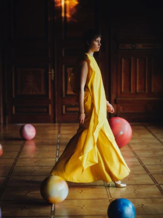 An adult female is walking in a yellow gown in an indoor space with large colorful spheres, wooden floors and panelling, and natural lighting.