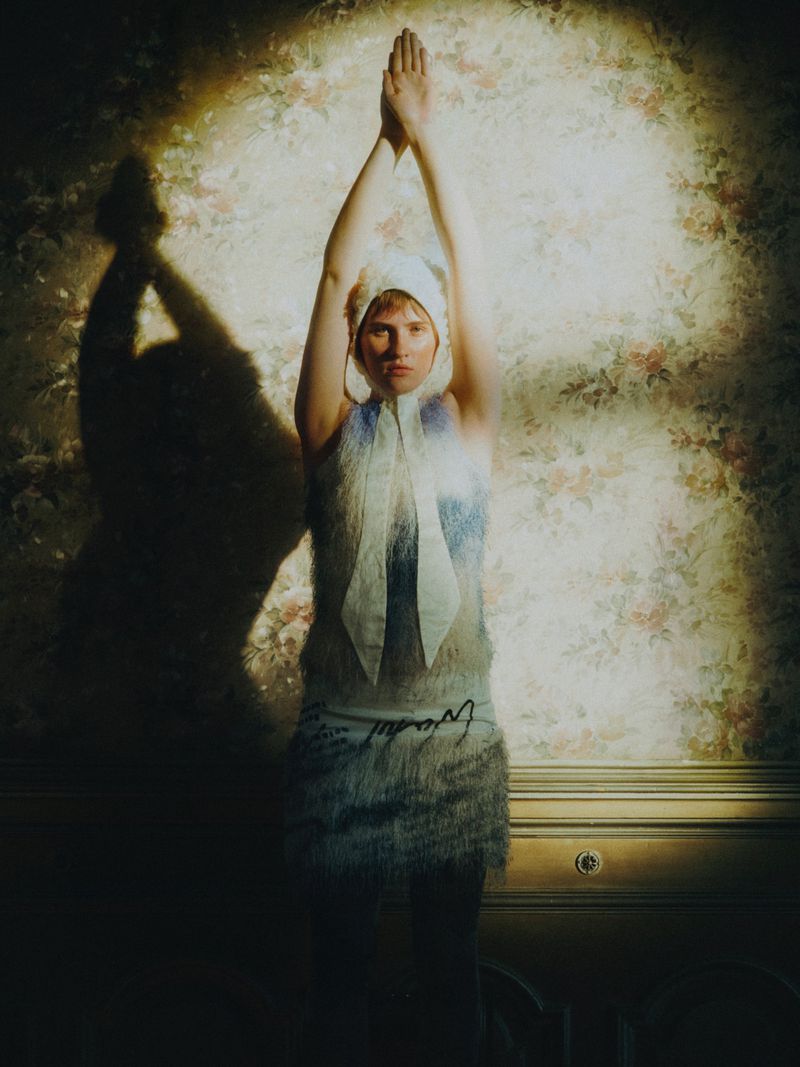 An adult female poses in a white dress with a floral background and soft lighting, hands raised above her head, casting a shadow.