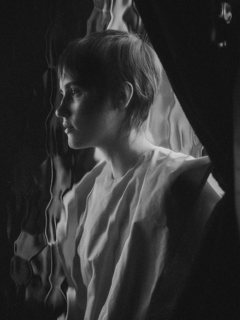 Monochromatic portrait of a young woman with short hair, in profile, wearing a light-colored top, framed by textured glass and shadows.