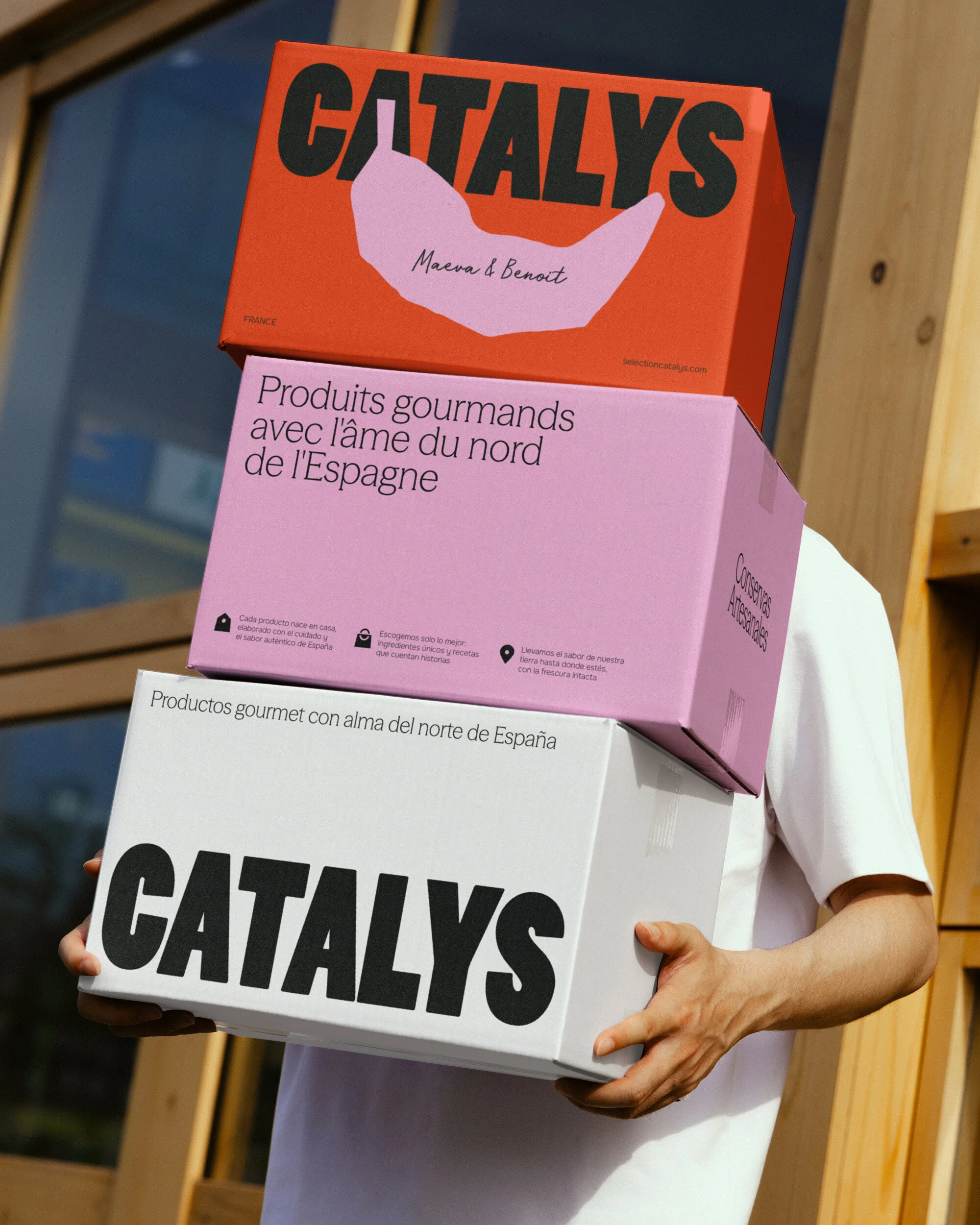 Person holds stacked branded Catalys boxes with gourmet food text in English, French, and Spanish, in an outdoor urban setting.