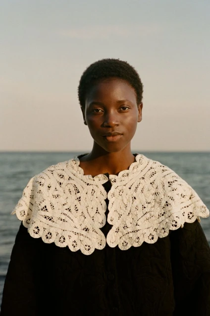 Portrait of a Black female model wearing a white lace collar against a blurred ocean background for a fashion editorial shoot.