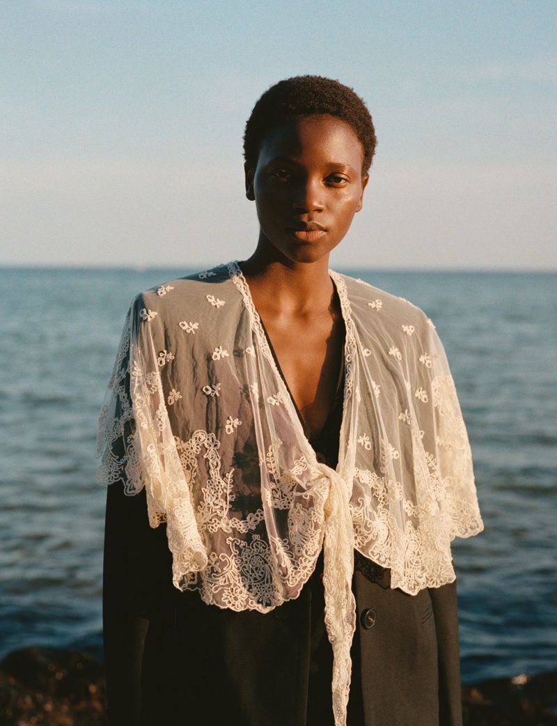 Woman in lace shawl poses against ocean backdrop in natural light portrait, emphasizing textures and serene mood.