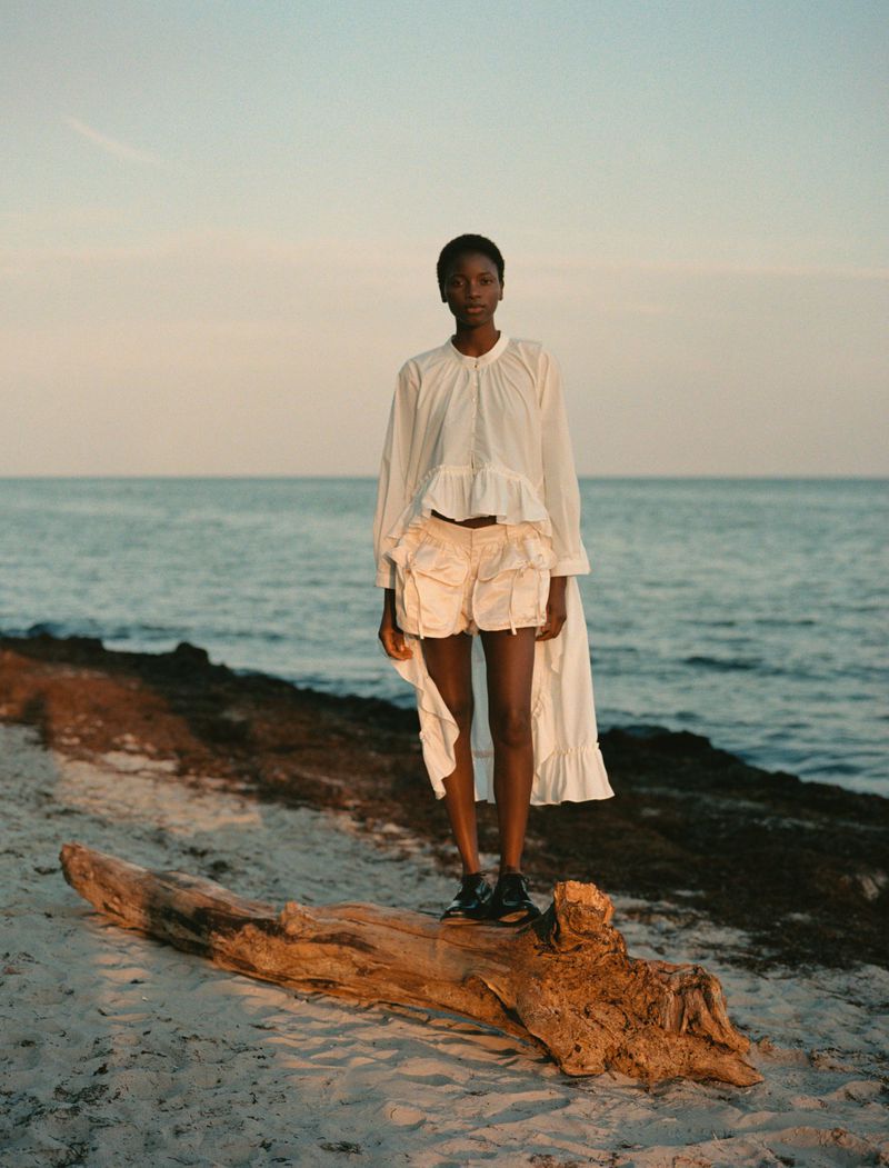 Woman in white blouse and shorts standing on a log at the beach with an ocean backdrop during golden hour.