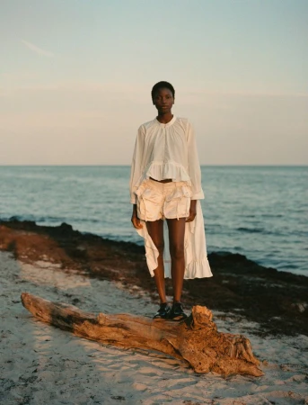 Woman in white blouse and shorts standing on a log at the beach with an ocean backdrop during golden hour.