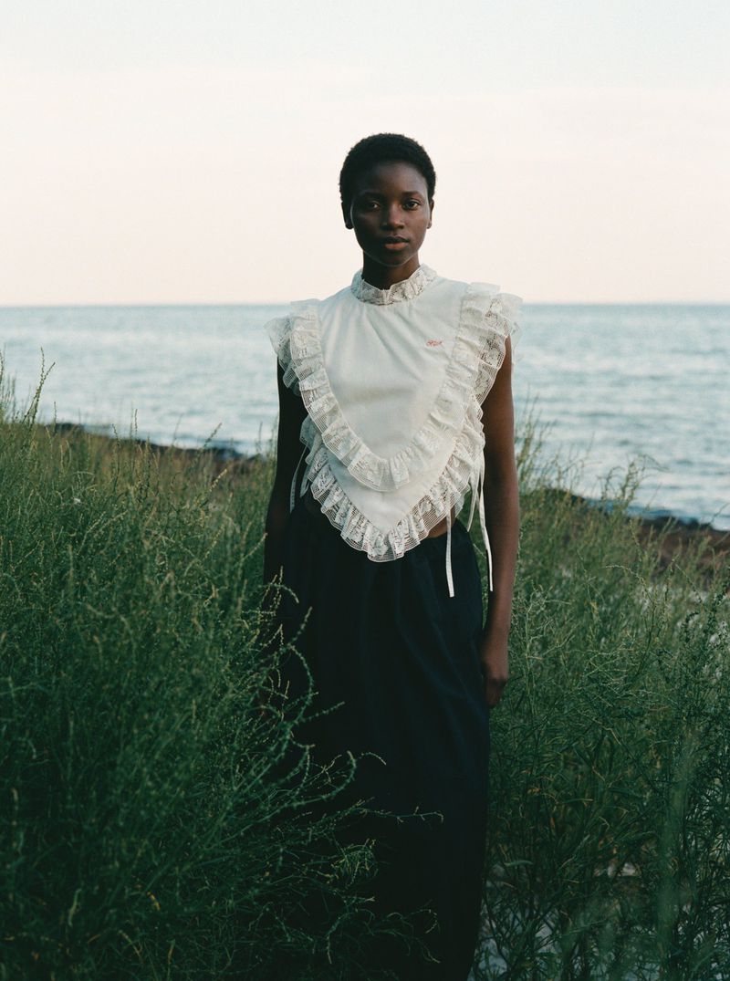 A solitary Black female model wearing a white ruffled top stands in tall grass against a pale ocean backdrop in a fashion editorial.