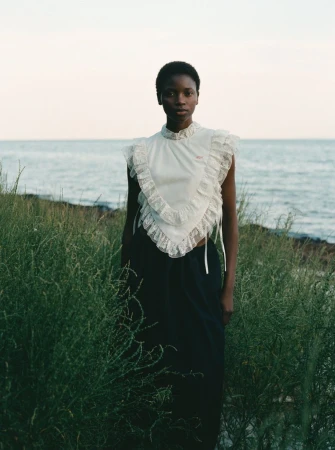 A solitary Black female model wearing a white ruffled top stands in tall grass against a pale ocean backdrop in a fashion editorial.