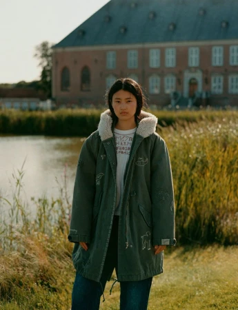 Young woman in an olive drab coat standing in a field with a pond and building in the background, photographed outdoors in natural light.