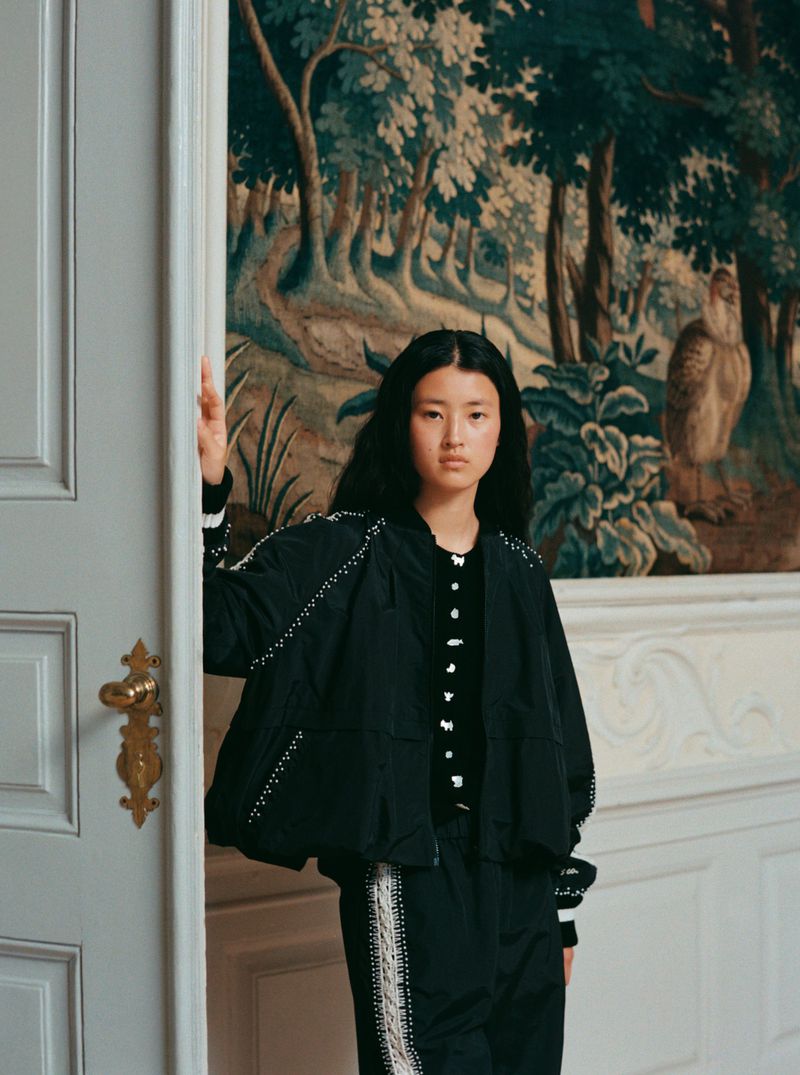 An Asian woman in a black jacket and pants stands in a doorway with a tapestry hanging on the wall in the background.