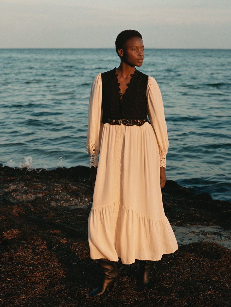 Woman poses in an off-white maxi dress with a black vest and boots on a rocky shore with the ocean in the background during daytime.
