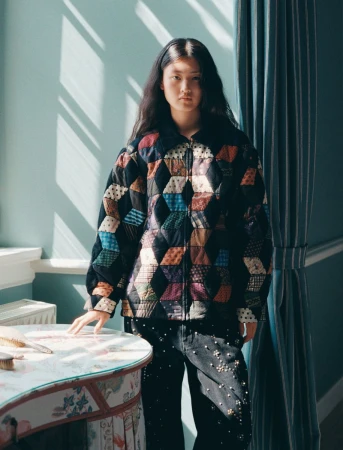 Portrait of a young woman with long dark hair wearing a colorful patchwork jacket and studded jeans, posing in front of a teal wall.