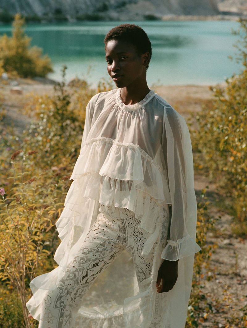 Elegant fashion portrait of an African woman in a white ruffled dress standing outdoors against a turquoise lake backdrop, natural light.