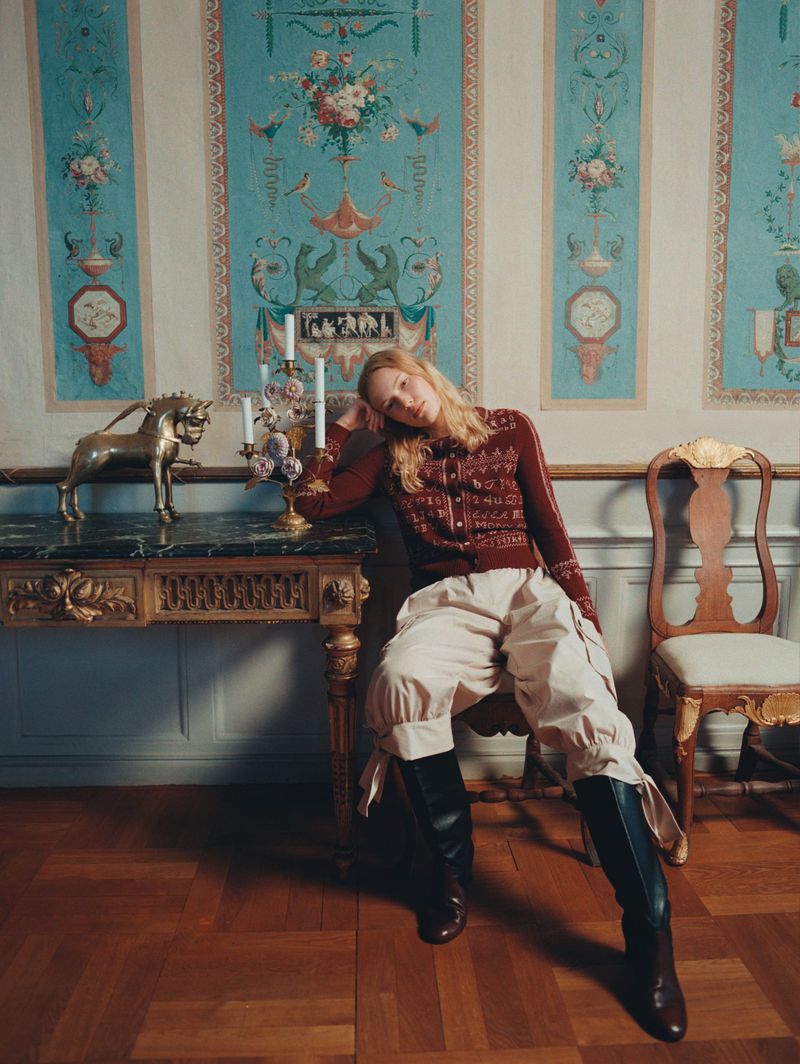 Indoor fashion shot of a woman posing in a cardigan, baggy pants, and boots, seated next to a table with decor in a designed interior.