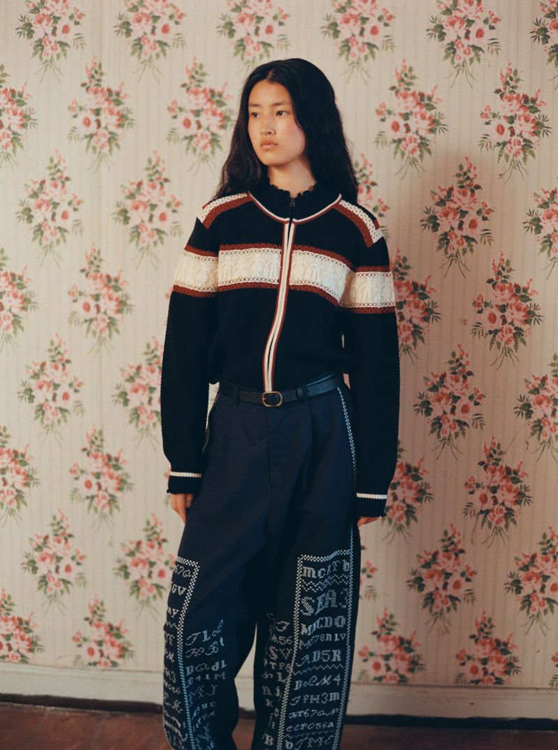 An Asian woman with long dark hair poses indoors, wearing a striped sweater and embroidered navy pants against a floral wallpaper.