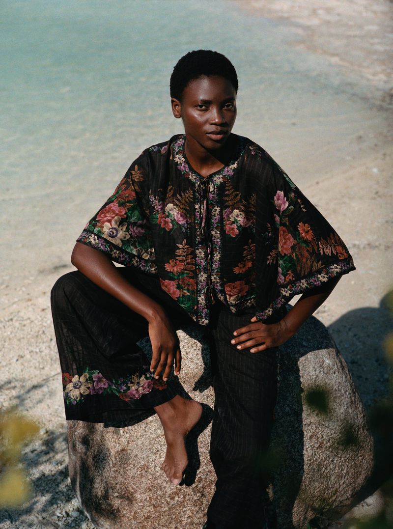 A black woman in a floral top and black pants sits on a rock at the beach in front of a body of water on a sunny day.