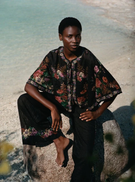 A black woman in a floral top and black pants sits on a rock at the beach in front of a body of water on a sunny day.
