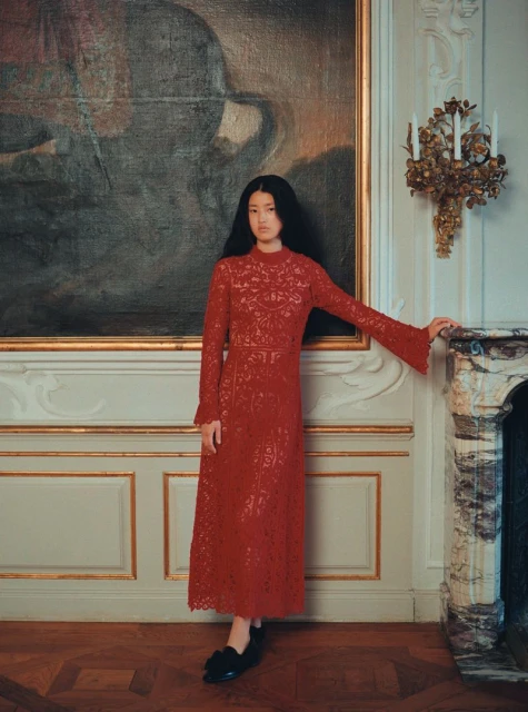 An elegant, full-length portrait of an Asian woman in a red lace dress, standing in front of a vintage painting in an ornate interior.