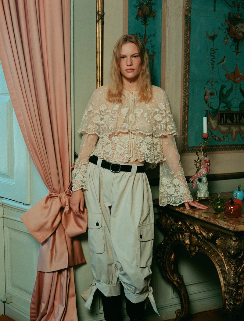 A person with blonde hair poses in a lace top and wide-legged cargo pants next to an ornate table, with a pink curtain in the background.