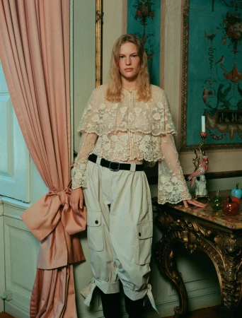 A person with blonde hair poses in a lace top and wide-legged cargo pants next to an ornate table, with a pink curtain in the background.