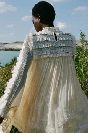 Model seen from behind wearing an ornate cream lace dress standing near water under a blue sky