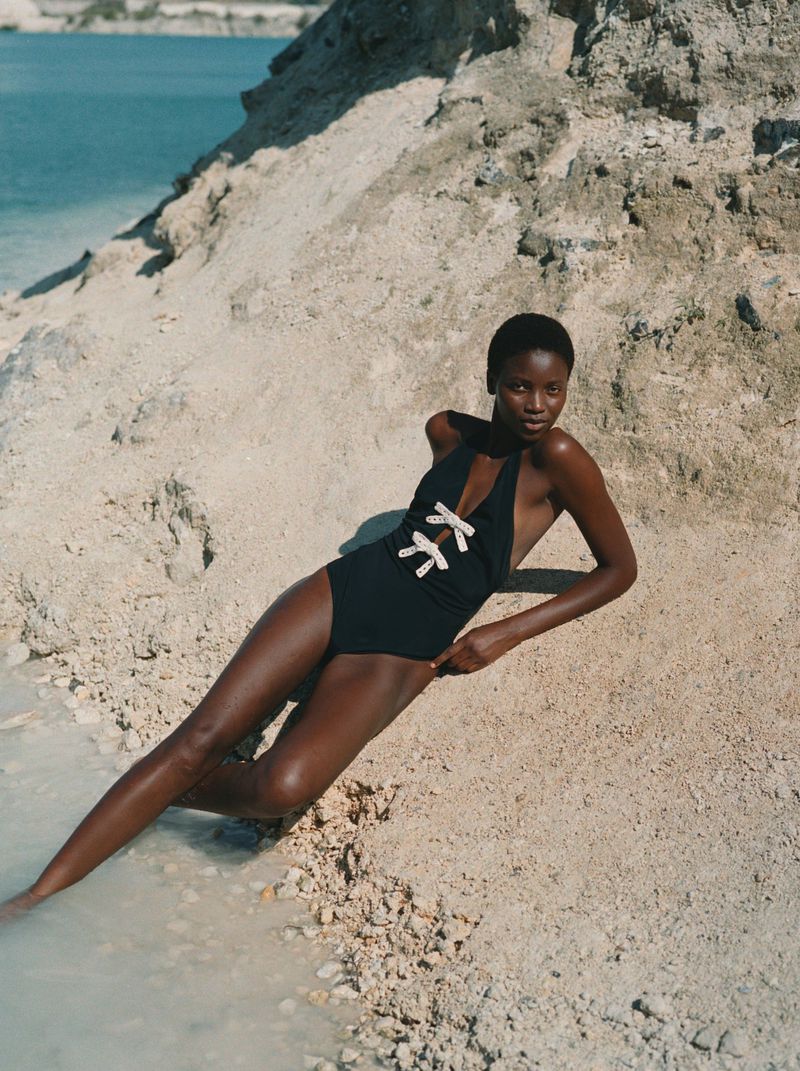 Woman in a black halter swimsuit lying on a sandy beach with turquoise water in the background, fashion editorial photograph.