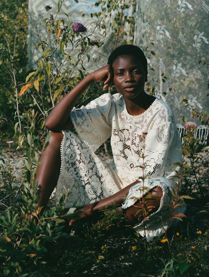 Young African woman in a lace dress sitting among lush greenery in soft, natural light in an outdoor setting, looking at the camera.