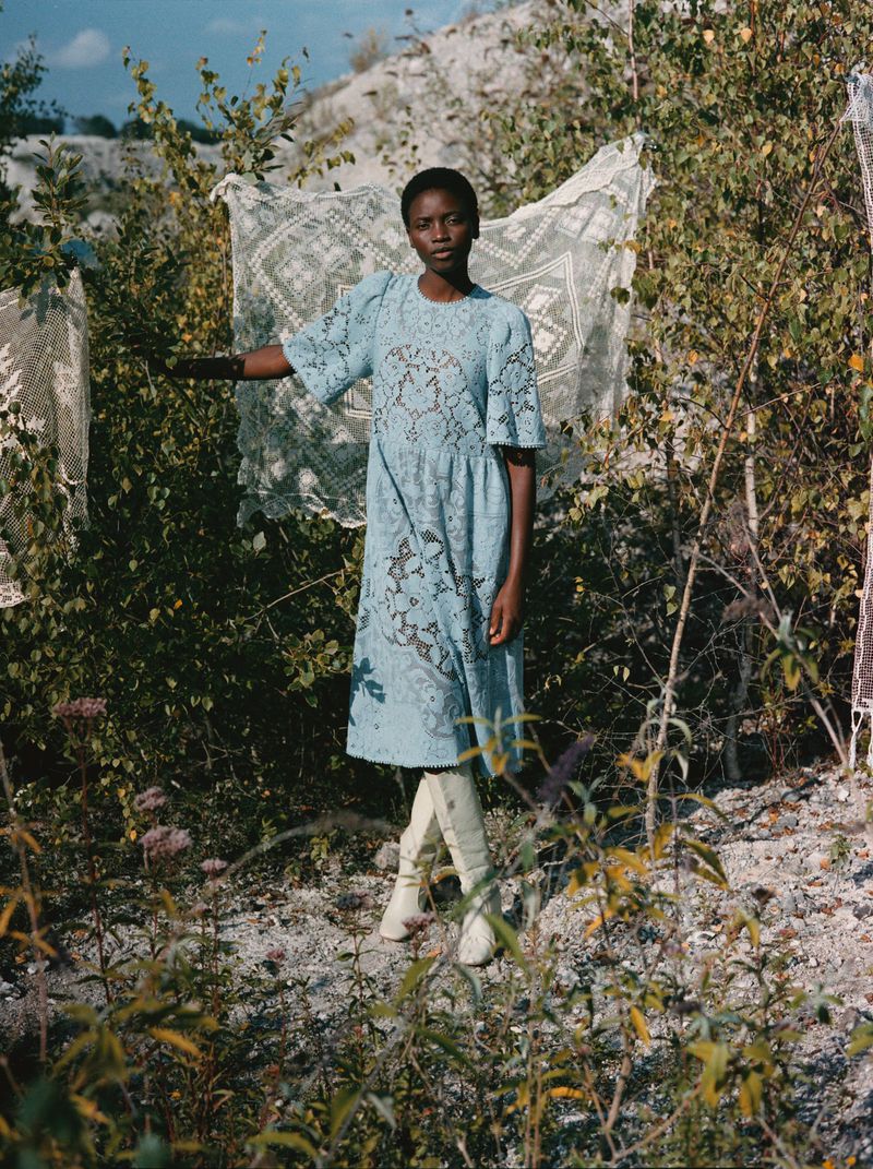 Woman in patterned dress outdoors near hanging fabrics, wearing boots. Bushes and stone gravel in foreground and a slope in background.