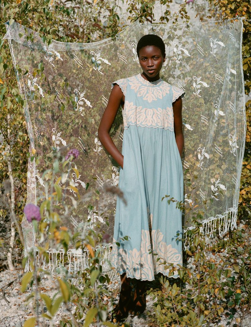 A dark-skinned woman in a light blue dress with lace detailing stands outside in natural light, framed by foliage and a lace curtain.