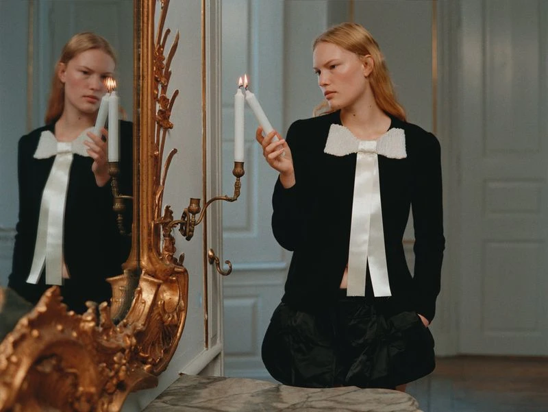 Woman in black jacket with bow poses indoors, holding candles and reflected in vintage gold mirror, fashion portrait, elegant indoor lighting.