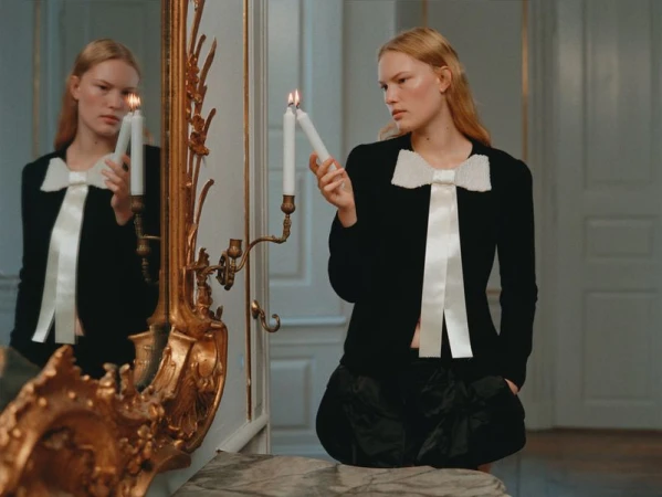Woman in black jacket with bow poses indoors, holding candles and reflected in vintage gold mirror, fashion portrait, elegant indoor lighting.