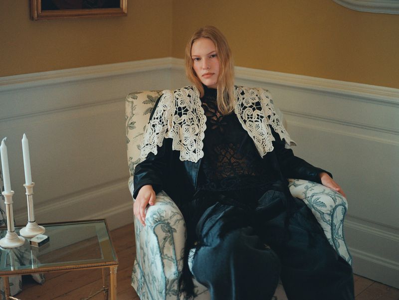 Portrait of a blonde adult woman seated in a patterned armchair in an interior setting with dark clothing and white lace collar.