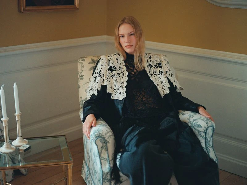 Portrait of a blonde adult woman seated in a patterned armchair in an interior setting with dark clothing and white lace collar.