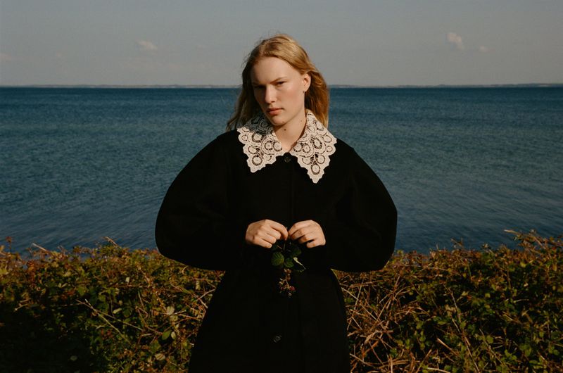Woman with blonde hair poses in a black coat with a white lace collar, holding greenery in a field by the sea under sunlight.