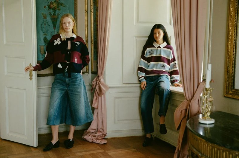 Two young women model striped rugby shirts and denim skirts in an elegant interior with hardwood floors, draped curtains, and ornate artwork.
