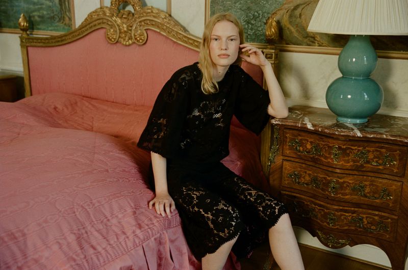 A fair-skinned woman in a black lace dress sits on a pink bed in a room with antique furniture and a painted mural backdrop.