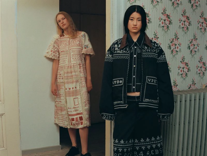 Two fashion models showcase embroidered dresses in an indoor setting with floral wallpaper, standing in a doorway with contrasting styles.
