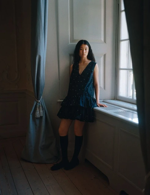 Asian woman in a black polka dot dress stands near a window, showcasing fashion in an indoor setting with soft, natural lighting.
