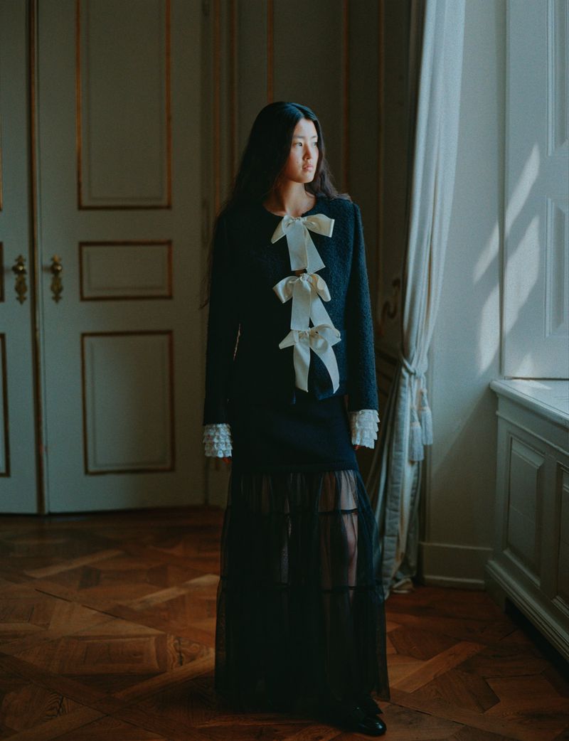 Elegant woman in dark skirt suit with white bow details poses beside a window in an ornately decorated room with hardwood floors.
