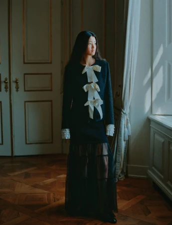 Elegant woman in dark skirt suit with white bow details poses beside a window in an ornately decorated room with hardwood floors.