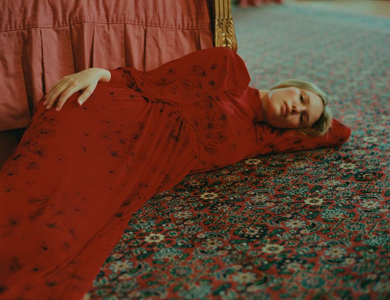 Woman in a long red patterned dress lies on an ornate rug in a brightly lit room with a bed visible in the background.