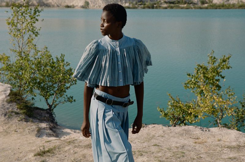 An African woman poses against a turquoise lake wearing a light blue pleated top and skirt, a black belt, and green foliage.