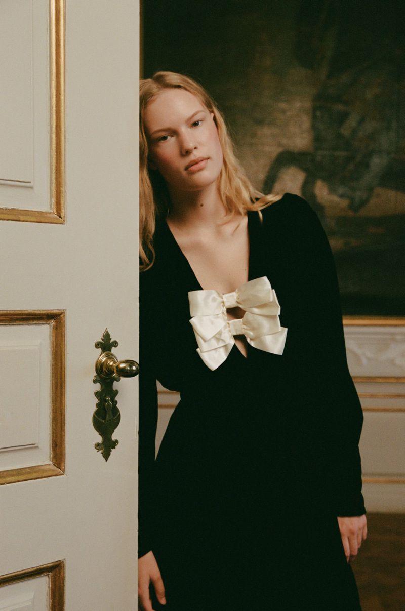 A fair-skinned adult woman in a black long-sleeve dress with large white bows stands near a white door, with a painting in the background.