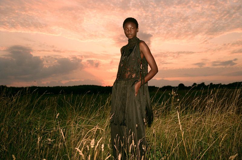 Woman in sleeveless dark dress standing in field of tall grass during sunset, looking at the camera in a fashion editorial.