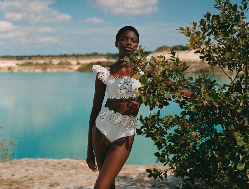 A striking photograph features a Black woman in a white two-piece outfit, posing by a leafy tree with a turquoise lake in the background.