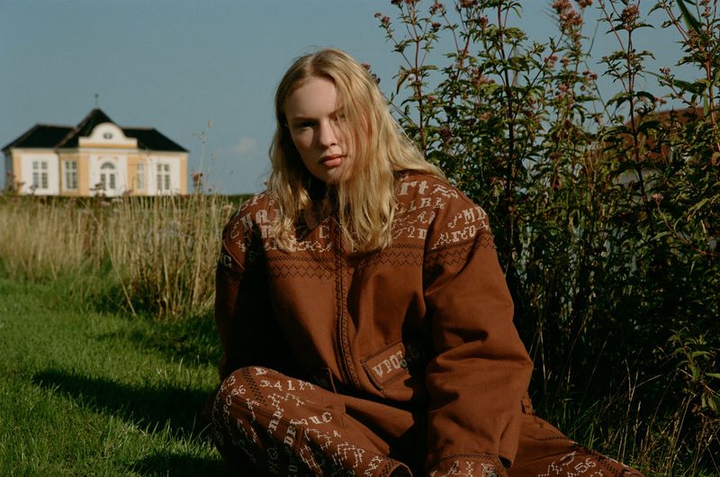 A blonde woman in a patterned brown jacket and pants sits on green grass in front of a field and a tan house under a blue sky.