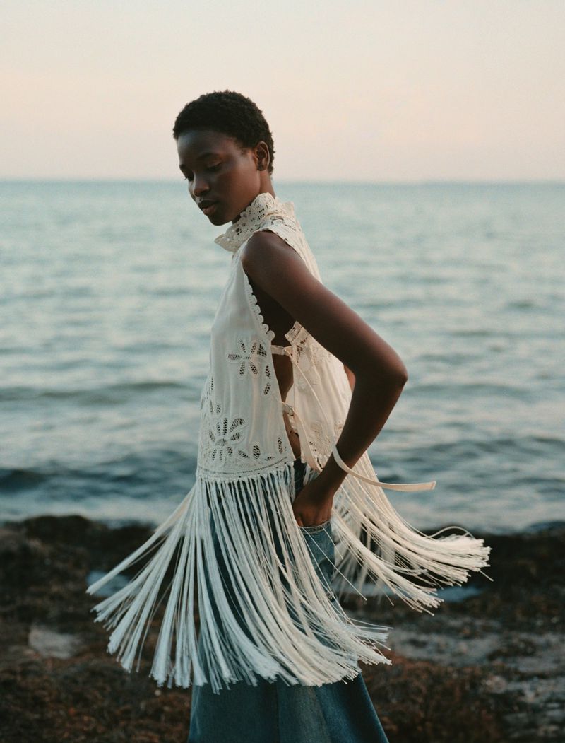 A woman stands near the ocean wearing a sleeveless lace top with a fringe hem and blue jeans in a fashion editorial shot.