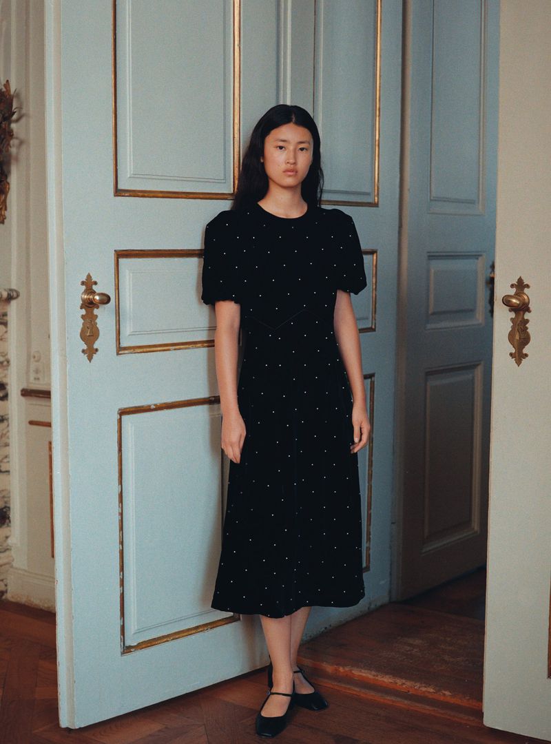 Elegant Asian woman in black midi dress with white polka dots posing near light blue door with gold trim in minimalist indoor setting.
