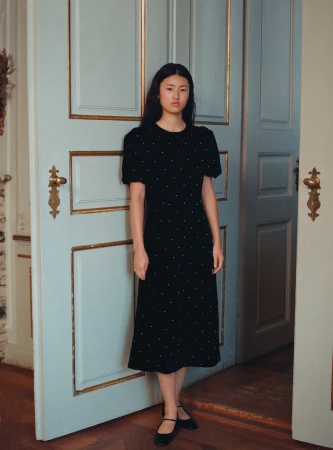 Elegant Asian woman in black midi dress with white polka dots posing near light blue door with gold trim in minimalist indoor setting.